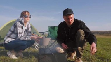 Orta yaşlı çift doğada pikniğin tadını çıkarıyor. Güneşli bir günde çadırınızın yanındaki portatif ızgarada mantar pişirmek. Mutlu erkek ve kadın dışarıda birlikte yemek pişiriyorlar.