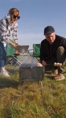 Genç bir adam ve kadın, güzel bir bahar günü dikey bir videoda kamp yaparken portatif bir kömür barbeküsünde taze yemek pişiriyorlar.