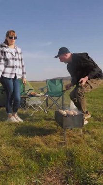 Genç bir adam ve kadın, güzel bir bahar günü dikey bir videoda kamp yaparken portatif bir kömür barbeküsünde taze yemek pişiriyorlar.