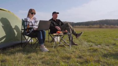 Dizüstü bilgisayarıyla uzaktan çalışan genç bir kadın kamp koltuğunda partnerinin yanında oturuyor güneşli bir günde açık havada dijital göçebe yaşam tarzının tadını çıkarıyor şişte barbekü mantarları yiyor..