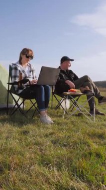 Yeşil bir alanda eşiyle kamp yaparken dizüstü bilgisayarda yazan serbest çalışan bir kadın dijital göçebe yaşam tarzını uzaktan çalışma ve açık hava macera videosuyla kucaklıyor.