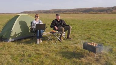Çiftler kamp yaparken güneşli günlerin tadını çıkarıyorlar. Kadın dizüstü bilgisayarda çalışıyor ve adam dijital göçebe yaşam tarzını gösteren küçük yemek masasının yanında dinleniyor.