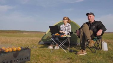 Yeşil çadırı olan kamp alanının keyfini çıkaran çift, bir kadın, bir serbest çalışan, dizüstü bilgisayarında çalışıyor ve partneri, açık hava barbeküsü için ızgarada patates kızartması yaparken yakınlarda dinleniyor.