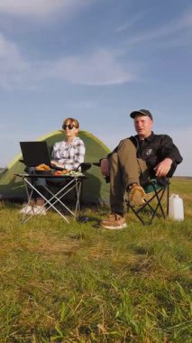 Yeşil bir alanda eşiyle kamp yaparken dizüstü bilgisayarda yazan serbest çalışan bir kadın dijital göçebe yaşam tarzını uzaktan çalışma ve açık hava macera videosuyla kucaklıyor.