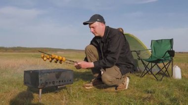 Adam güneşli bir kamp gününde şişte mantar kızartıyor. Karısıyla birlikte arka planda çadırlı yeşil bir alanda pikniğin tadını çıkarıyor.