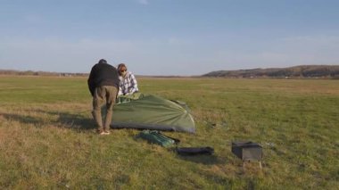 Birkaç gezgin, güneşli bir günde yeşil bir turist çadırını açmak ve bir araya getirmek için geniş bir açık alanda kamp yerlerini hazırlıyorlar.