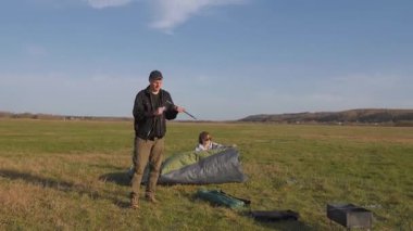 Birkaç gezgin, güneşli bir günde yeşil bir turist çadırını açmak ve bir araya getirmek için geniş bir açık alanda kamp yerlerini hazırlıyorlar.
