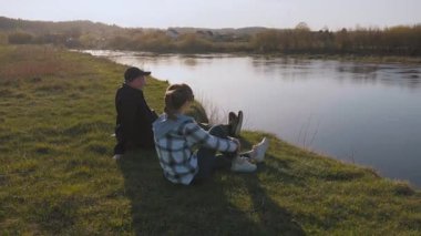 Olgun bir çift nehir kenarındaki yeşil çimlerde dinleniyor, manzaranın tadını çıkarıyor ve hafta sonu kaçamağı sırasında sakin bir açık hava yaşam tarzını somutlaştırıyorlar.