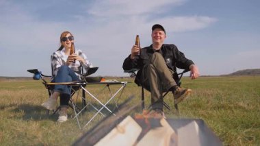 Genç adam ve kadın katlanır sandalyelerde oturur, engin yeşil bir tarlada barbeküyle pikniğin tadını çıkarırken, kadın bira içer ve güneşli bir günde birlikte dinlenir.
