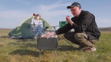 Genç adam ızgarada ızgara tavuk sosisi pişirirken kız arkadaşı da güneşli yeşil bir alanda çadırlarının yanında bira içip hafta sonu kaçamağının tadını çıkarıyor.