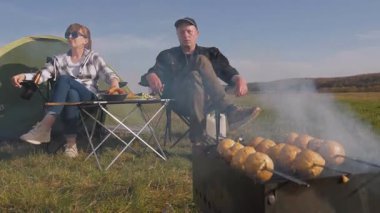 Genç bir adam ve kız, güneşli bir tarlada çadırlarının yanındaki kamp sandalyelerinde dinleniyor, mantar mantarlarını ızgaranın üstünde kızartıyorlar ve temiz havada huzurlu bir pikniğin tadını çıkarıyorlar.