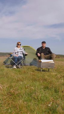Olgun bir adam barbekü için ızgara yaparken bir kadın arka plandaki dikey videoda gölle birlikte yeşil bir çayırda piknik yaparken çadırın yanındaki katlanır sandalyede dinleniyor.
