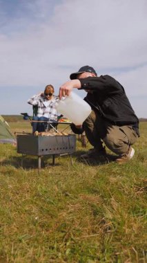 Genç bir adam barbeküde şiş kebap pişirirken kız arkadaşı da çadırlarının yanındaki çimenlik alanda kamp koltuğunda rahatlıyor ve birlikte pikniğin tadını çıkarıyor.