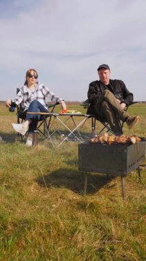 Mutlu çift kömür ızgarasında leziz ızgara yaparken manzaralı yeşil alanda piknik yaparken güneşli bir günde birlikte açık hava maceralarının keyfini çıkarırken ve kamp çadırlarının yanında dikey video görüntüleriyle birlikte.