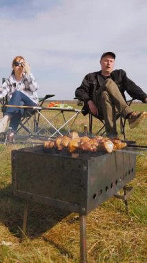 Beyzbol şapkalı genç bir adam güneşli bir günde arkadaşlarıyla açık hava pikniğinde şiş kebap pişirip şiş pişirip şiş kebap pişiriyor ve sıcak kömür üzerinde terbiye edilmiş et pişiriyor.