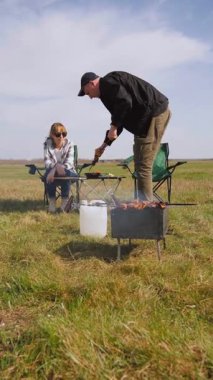 Beyzbol şapkalı genç bir adam güneşli bir günde arkadaşlarıyla açık hava pikniğinde şiş kebap pişirip şiş pişirip şiş kebap pişiriyor ve sıcak kömür üzerinde terbiye edilmiş et pişiriyor.