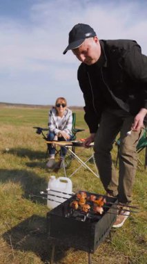 Beyzbol şapkalı genç bir adam güneşli bir günde arkadaşlarıyla açık hava pikniğinde şiş kebap pişirip şiş pişirip şiş kebap pişiriyor ve sıcak kömür üzerinde terbiye edilmiş et pişiriyor.