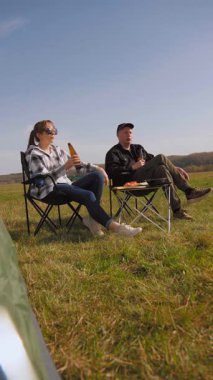 Genç çift kamp sandalyelerinde oturuyor, bira içiyor, konuşuyor ve güneşli bir günde güneşli bir alanda rahatlatıcı bir barbekü pikniğinin tadını çıkarıyor.