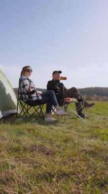 Mutlu bir adam ve kadın çadırlarının yanındaki kamp sandalyelerinde dinleniyor. Güneşli bir günde çimenli bir tarlada dikey bir videoda şiş kebabıyla barbekü pikniğinin keyfini çıkarıyorlar.