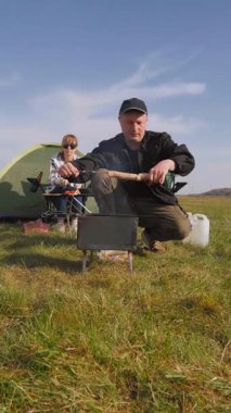 Mutlu yetişkin çift güneşli bir günde pikniğin keyfini çıkarırken, erkek barbeküde sosis pişirirken kadın güzel yeşil bir çayırda kamp masasında oturuyor.