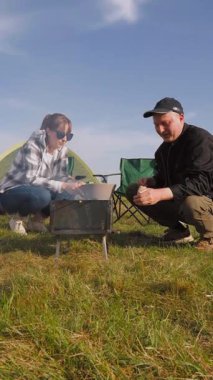 Mutlu bir adam ve kadın çadırlarının yanındaki kamp sandalyelerinde dinleniyor. Güneşli bir günde çimenli bir tarlada dikey bir videoda şiş kebabıyla barbekü pikniğinin keyfini çıkarıyorlar.