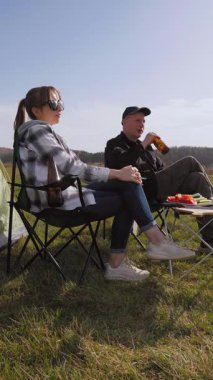 Mutlu bir adam ve kadın çadırlarının yanındaki kamp sandalyelerinde dinleniyor. Güneşli bir günde çimenli bir tarlada dikey bir videoda şiş kebabıyla barbekü pikniğinin keyfini çıkarıyorlar.