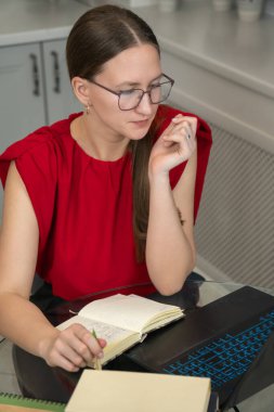 Kırmızı bluzlu kadın dizüstü bilgisayarla ilgileniyor, verileri analiz ediyor. O, kitaplarla çevrili, düşünceli bir ifadeye sahip parlak, modern bir odada cam bir masada oturuyor. Dikey fotoğraf