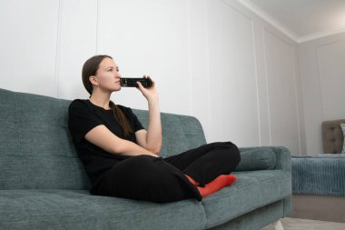 Siyah elbiseli ve kırmızı çoraplı genç bir kadın elinde uzaktan kumandayla bir kanepede oturuyor düşünceli ve sıkılmış bir halde televizyonda ne izleyeceğine karar vermeye çalışıyor.