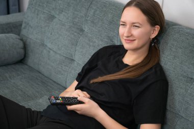 Mutlu genç kadın rahat bir kanepeye uzanıyor, uzaktan kumandayı tutuyor, komedi filmi veya TV programı izliyor, gülüyor ve eğleniyor, evde boş zamanlarının tadını çıkarıyor.