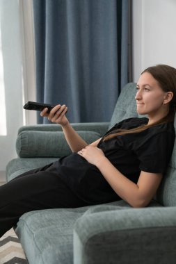 Comfortable young woman lounging on a sofa, watching television and holding a remote control, savoring a peaceful day at home filled with relaxation and leisure. Vertical photo
