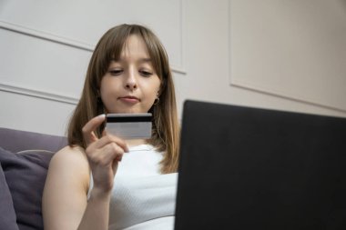 Kız, evdeki koltukta otururken dizüstü bilgisayar ve kredi kartıyla online ödeme yapıyor. Uygun ve güvenli e-ticaret deneyiminin tadını çıkarıyor.