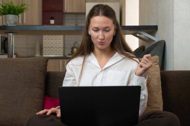 İş kadını kucağında dizüstü bilgisayarla kanepede oturuyor ve evde çalışırken şaşkın şaşkın bakıyor, kollarını kaldırıyor ve şok içinde ağzını açıyor.