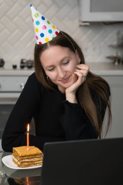 Parti şapkası takan genç bir kadın bilgisayarının önünde doğum günü pastası ve mumla doğum gününü internette kutluyor. Dikey fotoğraf