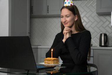 Doğum gününü internetten kutlayan genç bir kadın. Parti şapkası takıyor. Evde dizüstü bilgisayarının önünde yanan bir mumla dilek diliyor.