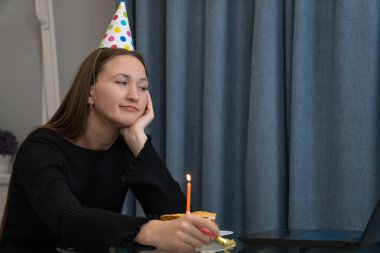 Parti şapkalı üzgün kadın doğum günü pastasına bakıyor ve elinde Noel kornosu tutuyor. Karantina sırasında evde tek başına kutlama yapıyor. Ailesiyle video görüşmesi yapıyor.