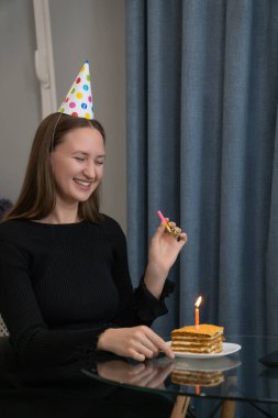 Parti şapkası takan gülümseyen genç bir kadın, önünde bir doğum günü pastası ve yanan bir mumla doğum gününü İnternet 'te kutlarken parti düdüğü çalıyor. Dikey fotoğraf