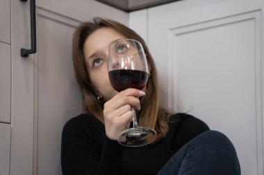 Düşünceli genç bir kadın mutfak zemininde oturmuş, elinde bir kadeh kırmızı şarapla yukarıya bakıyor, yalnızlığı ve etrafındaki derin düşünceleri somutlaştırıyor.