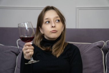 Kanepede oturan ve bardaktan kırmızı şarap içen, aroması ve tadı tadını çıkaran genç bir kadın, bir anlık rahatlama ile içeceğin nüanslarını düşünüyor.