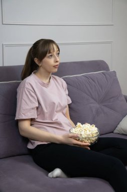 Sinema gecesinden zevk alan rahat bir kadın oturma odasında kanepede oturup ev eğlencesi yaşarken cam kaseden patlamış mısır yiyor. Dikey fotoğraf