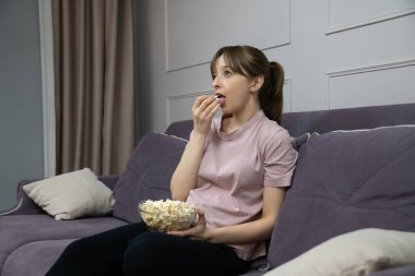 Gri bir kanepede rahat bir şekilde oturan genç bir kadın, oturma odasında büyüleyici bir film izlerken bir kase patlamış mısırın tadını çıkarıyor ve rahat ve keyifli bir atmosfer yaratıyor.