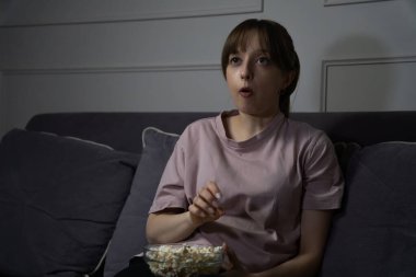 Şaşırmış genç bir kadın, geceleri oturma odasındaki rahat bir kanepede oturup cam kaseden patlamış mısır yerken TV 'de film izliyor.