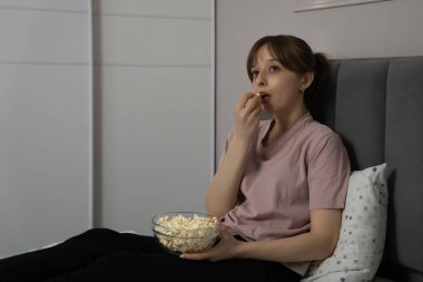 Genç kadın rahat bir şekilde yatağında oturmuş, büyüleyici bir film izlerken cam bir kaseden patlamış mısır yiyor, rahat ve eğlenceli bir ev sineması deneyimi yaratıyor.