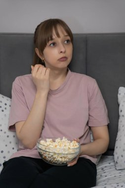 Genç bir kadın elinde patlamış mısırla yatağında oturmuş film seyrediyor. Şaşırmış ifadesi, planda beklenmedik bir değişiklik olduğunu gösteriyor. Dikey fotoğraf