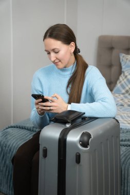 Bir otel odasında bavulun üzerinde oturan genç bir turist akıllı telefonunu kullanarak seyahat uygulamalarını kontrol ediyor ve heyecan verici bir gezi ve macera planlarını tamamlıyor. Dikey fotoğraf