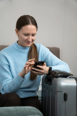 Genç bir kadın bavulunun yanında yatarken akıllı telefon kullanıyor, seyahat planlıyor ve online rezervasyon yaptırıyor ya da seyahat bilgilerini kontrol ediyor. Dikey fotoğraf