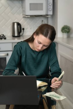 Genç bir kadın ev bütçesini yönetiyor, Amerikan dolarlarını sayıyor ve mutfak masasında oturmuş kişisel finans ve harcamalara odaklanırken dizüstü bilgisayar üzerinde çalışıyor. Dikey fotoğraf