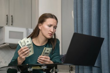 Ev bütçesini yöneten genç bir kadın dizüstü bilgisayar kullanırken dikkatle dolarları sayıyor. Muhtemelen online bankacılık ya da masraf takibi için.