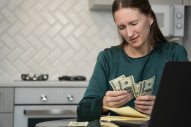 Genç bir kadın ev ekonomisini yönetiyor, kalan dolarları sayıyor, dizüstü bilgisayarla mutfak masasında oturuyor, maddi sorunlar yüzünden stresli hissediyor ve faturaları ödemekte zorlanıyor.