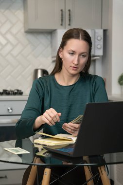 Ev ekonomisini yöneten, nakit para sayan ve dizüstü bilgisayar kullanan, mutfaktaki cam masada oturan, bütçeyi yöneten ve online bankacılık yapan genç bir kadın. Dikey fotoğraf