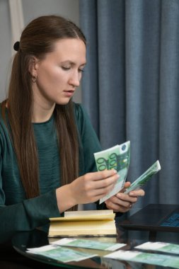 Genç kadın muhasebeci dikkatlice avro banknotlarını sayıyor, finans yönetiyor, bütçeyi hesaplıyor ve dizüstü bilgisayarla not tutuyor. Dikey fotoğraf